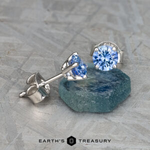 The EA-DB-402 earrings elegantly feature medium blue sapphires, each gemstone adding a sophisticated touch, beautifully set on a textured silver surface.