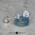The EA-DB-401 earrings, adorned with sparkling light blue sapphires, are showcased on a rough blue stone. The textured light gray background enhances the depth and contrast, highlighting the refined elegance of these stunning accessories.