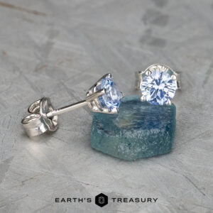 The EA-DB-401 earrings, adorned with sparkling light blue sapphires, are showcased on a rough blue stone. The textured light gray background enhances the depth and contrast, highlighting the refined elegance of these stunning accessories.