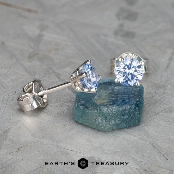 EA-DB-401 The EA-DB-401 earrings, adorned with sparkling light blue sapphires, are showcased on a rough blue stone. The textured light gray background enhances the depth and contrast, highlighting the refined elegance of these stunning accessories.