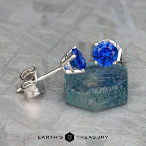 The EA-DB-403 earrings are a stunning pair featuring blue sapphire gemstones measuring 3.5-5.0 mm, set in silver, elegantly displayed on a textured surface and teal block.
