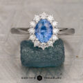 The "Cassandra" ring in 14K white gold, featuring a 1.42-carat Ceylon sapphire surrounded by a halo of clear stones, rests on a rough blue-green crystal above patterned metal. The Earth's Treasury logo appears below.