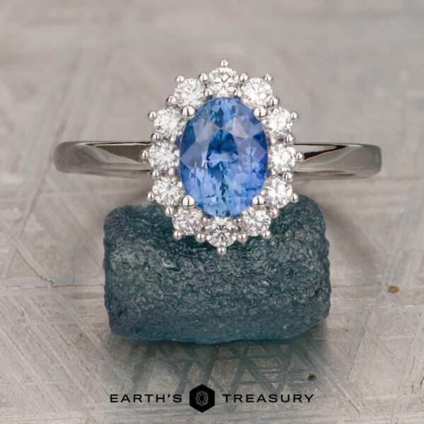 The "Cassandra" ring in 14K white gold with 1.42-carat Ceylon sapphire The "Cassandra" ring in 14K white gold, featuring a 1.42-carat Ceylon sapphire surrounded by a halo of clear stones, rests on a rough blue-green crystal above patterned metal. The Earth's Treasury logo appears below.