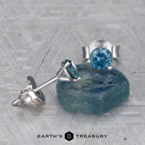 A pair of EA-MS-300 4.0mm Teal Martini Style silver earrings—one upright, one lying down on rough blue mineral—displayed on a textured gray surface with the "Earth's Treasury" logo at the bottom.