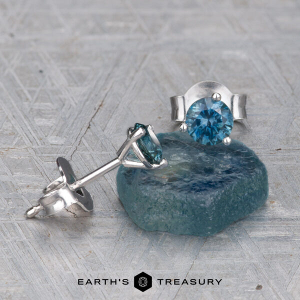 EA-MS-300 4.0mm Teal A pair of EA-MS-300 4.0mm Teal Martini Style silver earrings—one upright, one lying down on rough blue mineral—displayed on a textured gray surface with the "Earth's Treasury" logo at the bottom.