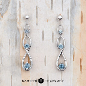 The EA-MS-601 earrings are elegant white gold drops with a twisting design, each set with three round Montana sapphires, shown on light wood with the Earth’s Treasury logo at the bottom.
