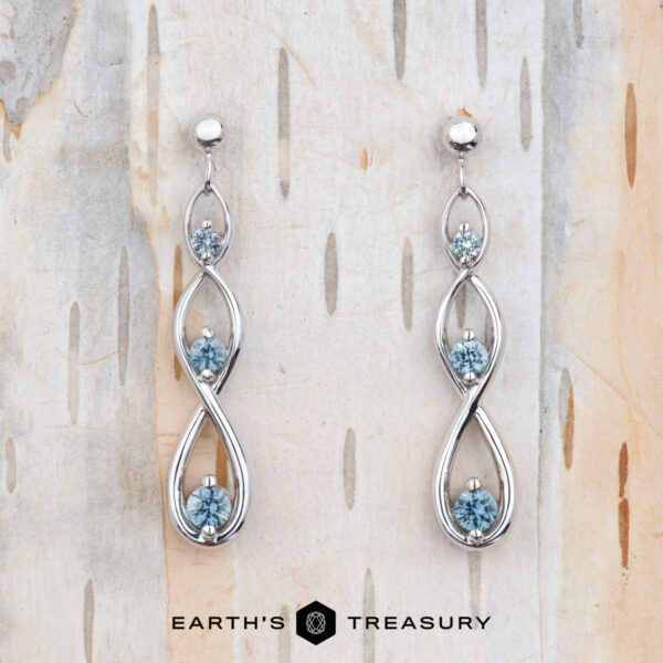 The EA-MS-601 earrings are elegant white gold drops with a twisting design, each set with three round Montana sapphires, shown on light wood with the Earth’s Treasury logo at the bottom.