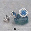 A pair of Montana Sapphire Diamond Halo Earrings, Medium Blue 4.5 mm (Heat), featuring round sapphires with diamond halos—one upright, the other resting on a rough blue stone.