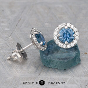 A pair of Montana Sapphire Diamond Halo Earrings, Medium Blue 4.5 mm (Heat), featuring round sapphires with diamond halos—one upright, the other resting on a rough blue stone.