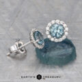 A pair of Montana Sapphire Diamond Halo Earrings, Light Aqua Blue 4.5 mm, in white metal settings—one upright on a blue stone, the other lying flat on a textured surface—with the Earth's Treasury logo below.
