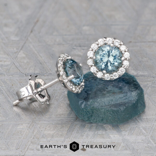 Montana Sapphire Diamond Halo Earrings, Light Aqua Blue 4.5 mm ( A pair of Montana Sapphire Diamond Halo Earrings, Light Aqua Blue 4.5 mm, in white metal settings—one upright on a blue stone, the other lying flat on a textured surface—with the Earth's Treasury logo below.