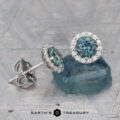 A pair of Teal Blue 4.5 mm Montana Sapphire Diamond Halo Earrings (Heated)—one flat, one upright—are displayed on a blue stone slab.