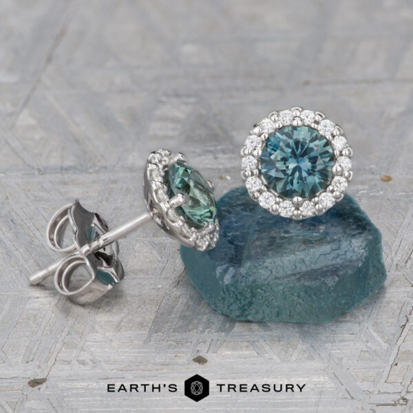 Montana Sapphire Diamond Halo Earrings, Teal Blue 4.5 mm (Heated A pair of Teal Blue 4.5 mm Montana Sapphire Diamond Halo Earrings (Heated)—one flat, one upright—are displayed on a blue stone slab.