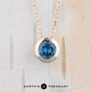 The FJ-NEC-101 4.5mm gold bezel slide necklace features a round teal blue gemstone pendant, showcased against a light, textured background. "EARTH'S TREASURY" is displayed at the bottom.