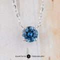 The FJ-NEC-105 5.0mm necklace features a round teal blue gemstone pendant in a prong station setting, suspended from a silver chain with the "Earth's Treasury" logo displayed at the bottom against a light background.