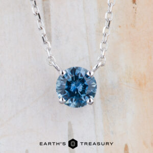 The FJ-NEC-105 5.0mm necklace features a round teal blue gemstone pendant in a prong station setting, suspended from a silver chain with the "Earth's Treasury" logo displayed at the bottom against a light background.