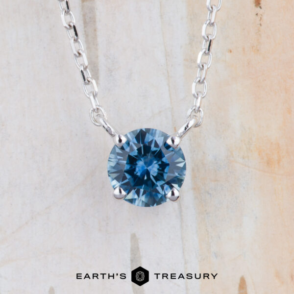 The "Glow" Prong Station Necklace - Teal Blue, 5.0 mm (Heated) The FJ-NEC-105 5.0mm necklace features a round teal blue gemstone pendant in a prong station setting, suspended from a silver chain with the "Earth's Treasury" logo displayed at the bottom against a light background.