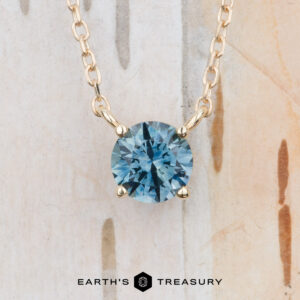 The "Glow" necklace in 14K yellow gold features a round teal blue gemstone pendant on a gold chain, displayed against a light textured background with the "Earth's Treasury" logo at the bottom.