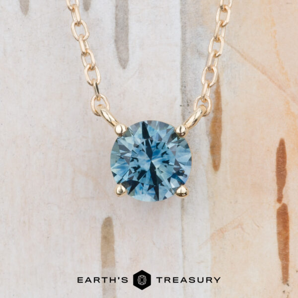 The "Glow" necklace in 14K yellow gold The "Glow" necklace in 14K yellow gold features a round teal blue gemstone pendant on a gold chain, displayed against a light textured background with the "Earth's Treasury" logo at the bottom.