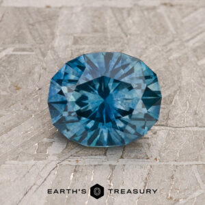 A 1.36-carat heated teal blue Montana Sapphire is displayed on a textured gray surface, with "EARTH'S TREASURY" written below.