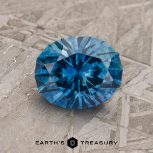 A 1.93-carat rich blue Montana Sapphire (heated), round and faceted, is displayed on a textured gray surface with "EARTH'S TREASURY" and a hexagonal logo beneath the stone.