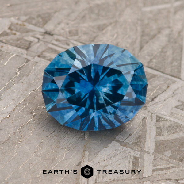 1.93-Carat Rich Blue Montana Sapphire (Heated) A 1.93-carat rich blue Montana Sapphire (heated), round and faceted, is displayed on a textured gray surface with "EARTH'S TREASURY" and a hexagonal logo beneath the stone.