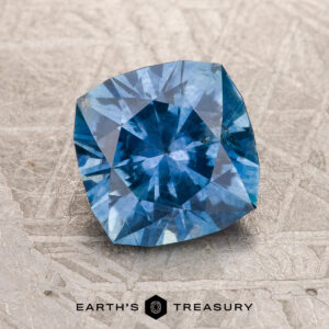 A 1.78-carat medium blue heated Montana Sapphire with a cushion cut rests on a textured gray surface, "EARTH'S TREASURY" displayed below.