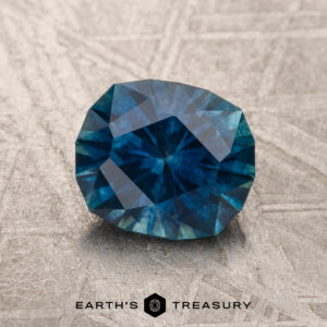 A 1.70-carat deep peacock blue Montana Sapphire (heated) rests on a textured metallic surface, with the "Earth's Treasury" text and logo displayed below.