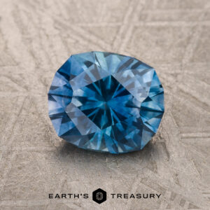 A 1.67-carat medium blue heated Montana Sapphire is displayed on a textured surface with the Earth's Treasury logo and name at the bottom.