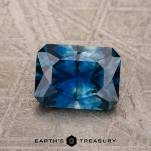 A 1.53-carat Deep Peacock Blue Montana Sapphire (Heated) with geometric facets rests on a textured surface, with the "Earth's Treasury" logo featured below.