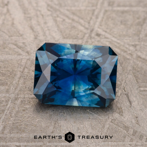 1.53-Carat Deep Peacock Blue Montana Sapphire (Heated) A 1.53-carat Deep Peacock Blue Montana Sapphire (Heated) with geometric facets rests on a textured surface, with the "Earth's Treasury" logo featured below.