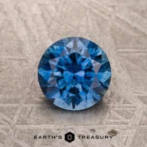 A 1.28-carat medium blue heated Montana Sapphire is shown, round and faceted, on a gray textured surface with “Earth’s Treasury” and its logo below.