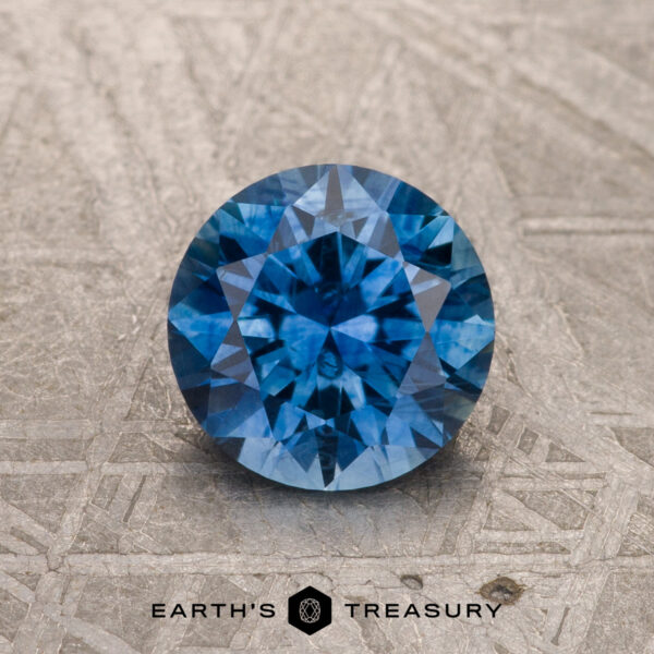 A 1.28-carat medium blue heated Montana Sapphire is shown, round and faceted, on a gray textured surface with “Earth’s Treasury” and its logo below.