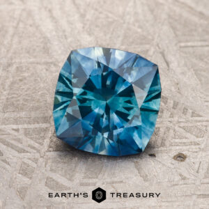 A 1.66-carat blue-teal particolored Montana Sapphire (heated), cushion-cut with intricate facets, rests on a textured gray surface above the Earth's Treasury logo.