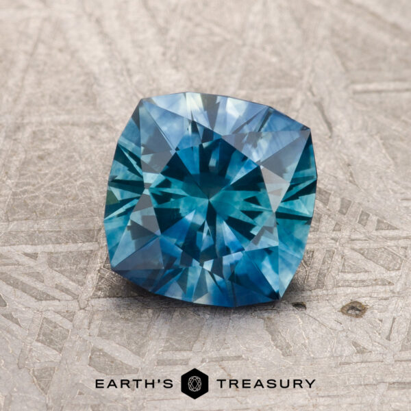 1.66-Carat Blue-Teal Particolored Montana Sapphire (Heated) A 1.66-carat blue-teal particolored Montana Sapphire (heated), cushion-cut with intricate facets, rests on a textured gray surface above the Earth's Treasury logo.