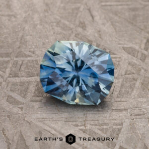A 1.27-carat blue-aqua particolored Montana Sapphire with a round, geometric cut is displayed on a textured gray surface, featuring the text "Earth's Treasury" at the bottom.