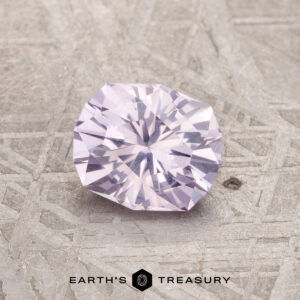 A round, faceted 1.16-carat pale pink Montana Sapphire rests on a textured metallic surface, with the "Earth's Treasury" logo and text below.