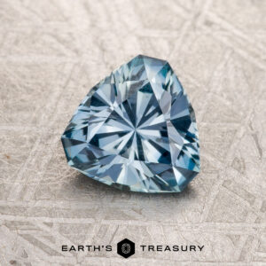 A 1.25-Carat Light Aqua Blue Montana Sapphire, trillion-cut and faceted, is displayed on a textured surface with the "Earth's Treasury" logo and text beneath it.