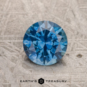A 1.31-carat medium blue heated Montana Sapphire is shown on a textured gray surface with "EARTH'S TREASURY" below.