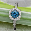 The “Paeonia” Ring features a 1.68-carat round Montana Sapphire in platinum, inspired by the Paeonia flower and accented with marquise and round clear stones, displayed on a green and white leaf with The Earth's Treasury logo below.