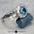 The “Paeonia” Ring in platinum, featuring a 1.68-carat Montana Sapphire, is displayed beside a rough blue stone on a textured surface. The text reads "Earth's Treasury.