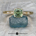 The “Melanie” ring in 18k yellow gold, featuring a 1.53-carat Australian sapphire and side diamonds, is displayed on a blue rough stone with the "Earth's Treasury" logo below.
