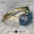 The “Melanie” ring in 18k yellow gold, featuring a 1.53-carat Australian sapphire and diamond accents, is displayed beside a rough blue stone on a gray textured surface. The "Earth’s Treasury" logo appears at the bottom.