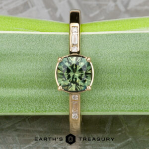 The “Melanie” ring in 18k yellow gold features a 1.53-carat Australian sapphire center, accented with four diamonds on the band. Shown vertically on green with the Earth's Treasury logo below, it’s a striking choice for any occasion.