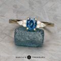 An Estes, Kayley and Nore, Caroline 1 ring with an oval blue gemstone and two clear side stones rests on a rough blue mineral, with the “Earth’s Treasury” logo below.