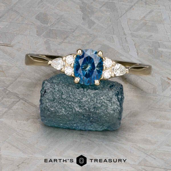 The "Arwyn" ring in 14K yellow gold with 0.95-carat Montana sapphire An Estes, Kayley and Nore, Caroline 1 ring with an oval blue gemstone and two clear side stones rests on a rough blue mineral, with the “Earth’s Treasury” logo below.