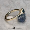 The Estes, Kayley and Nore, Caroline 1 ring, featuring a gold band with a blue gemstone and small diamonds, rests on a rough blue stone. The background is gray with a geometric pattern and “EARTH’S TREASURY” at the bottom.