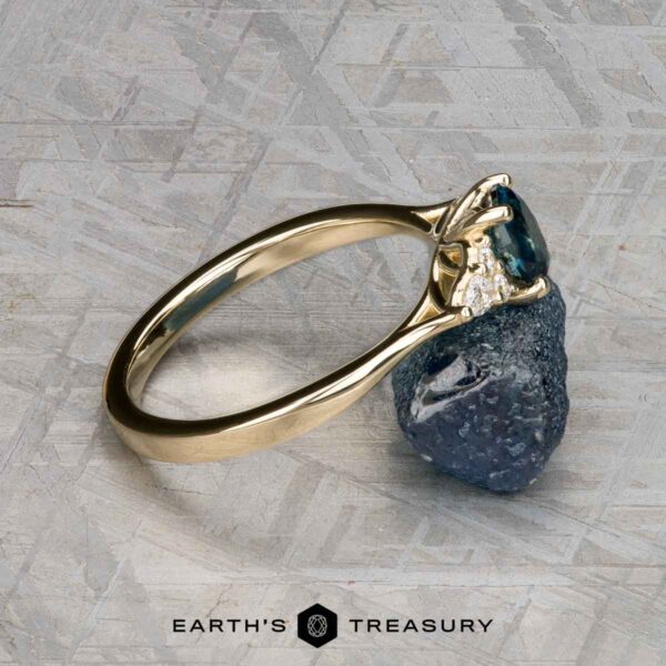 The "Arwyn" ring in 14K yellow gold with 0.95-carat Montana sapphire The Estes, Kayley and Nore, Caroline 1 ring, featuring a gold band with a blue gemstone and small diamonds, rests on a rough blue stone. The background is gray with a geometric pattern and “EARTH’S TREASURY” at the bottom.