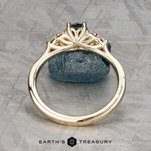 The Estes, Kayley and Nore, Caroline 1 ring features an intricate gold trellis cluster design around a dark gemstone, displayed upright on a textured gray surface with "EARTH'S TREASURY" at the bottom.