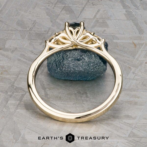 The "Arwyn" ring in 14K yellow gold with 0.95-carat Montana sapphire The Estes, Kayley and Nore, Caroline 1 ring features an intricate gold trellis cluster design around a dark gemstone, displayed upright on a textured gray surface with "EARTH'S TREASURY" at the bottom.